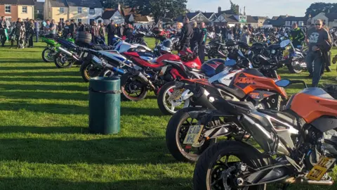 Lots of motorcycles parked on a village green, with lots of people walking about and talking.