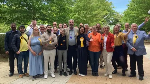 Emma Howgego/BBC Liberal Democrat councillors and members celebrate victory in South Cambridgeshire District Council in a group photo outside with trees in the background.