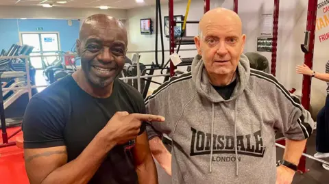 Two men standing in a gym. Left, the man is in a black t-shirt, smiling and pointing at the man on the right. The man on the right is wearing a grey hoodie with his hands on his hips.