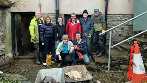 Eight people are posing in front of Yew Tree Hall. They are a little muddy after working on the redevelopment.