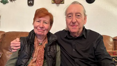 A woman with short red hair and man with long silver hair sit on a sofa smiling at the camera, the man has his arm around the woman's shoulder