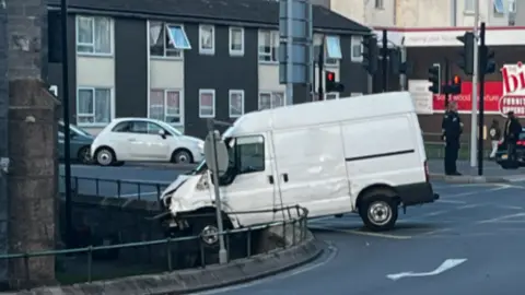 A picture of a large white van which has crashed into a roundabout. It has damaged the railings and police are pictured in the background.
