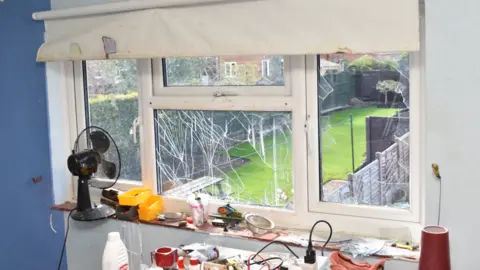 Bedfordshire Police A smashed window inside the house with chemicals on a table. You can see a garden, with green grass, a fan, and plugs. 