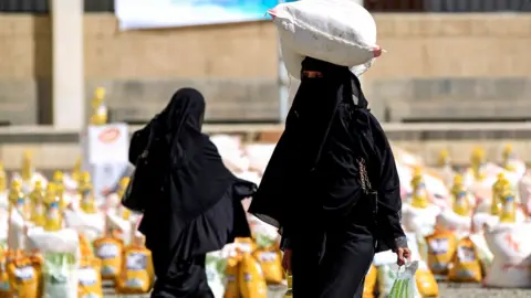 Getty images a woman carry a sack of grain on her head in black. There are lots of other sacks in the background.