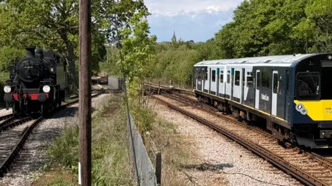 Jennie A black steam locomotive on the left of the image on a track with a modern electric train on the right on a track.