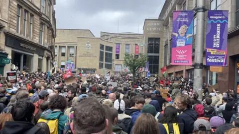 A large crowd fills a city street during a demonstration, with people holding placards and flags, facing toward a civic building, while surrounding shopfronts and banners—including ones reading “John Lewis” and “Glasgow Life”—are visible under overcast skies.