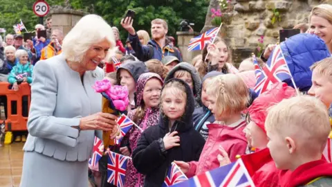 A beaming Queen Camila outside the gates of the abbey in a grey suit greeting a crown of children in colourful coats in the rain. Many are waving the Union Jack and there are adults in the background taking photos on their phones. 