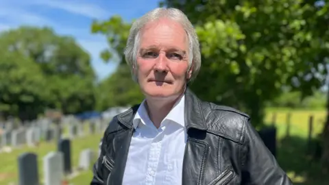 A man with grey hair, a white shirt and a black leather jacket is looking at the camera with a neutral expression. He is standing in a cemetery - behind him are rows of graves on a bright sunny day. 