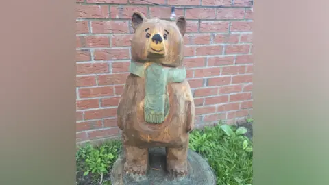 A small wooden carved bear sculpture on a circular wooden platform in a garden. The bear is brown and standing on its hind legs, smiling. It has a green scarf on