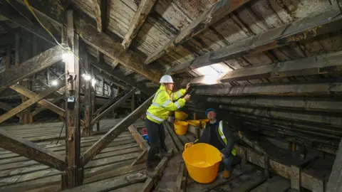 Pete Seaward Two builders in yellow hi vis jackets work on the inside of Blenheim Palace's roof. They have several yellow buckets with them. One of them is shining a flashlight at the rood while standing up. The other one is sitting below and looking at the light.