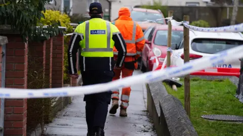 PA Media Garda walking down a path with another man in all orange uniform in front. Blue and white police tape is in the foreground, to the right of both men are parked cars, the nearest is white and the one further away is red.