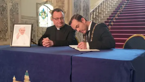 The book of condolence is being signed by the mayor with his gold sash on. Beside him is a priest wearing a black suit and white priest collar. The book and a picture of the pope is laid on a blue table with a blue table cloth featuring a Belfast City Hall print on it. 