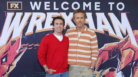 Wrexham co-chairmen and Executive Producers Rob Mac and Ryan Reynolds smile in front of branding for their documentary series "Welcome to Wrexham" at a red carpet event