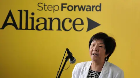 Pacemaker Anna Lo at an Alliance Party manifesto launch.  She is wearing a white jacket, a monochrome patterned top and is speaking into a microphone.