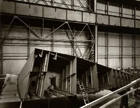 Bruce Rae A black and white photograph of a man inside a metal structure which looks to be part of a ship. It is in the process of being built and is on its side facing the camera. The man is dressed in black and looking into the camera. There is a ladder leaning against it. The structure looks to be inside a large warehouse with metal walls.