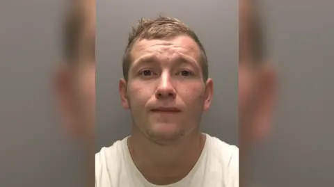 A police headshot of a man looking at the camera wearing a white t-shirt. He has short light-coloured hair and has brown eyes. 