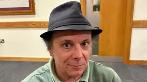 Martin Heath/BBC Ben Foley wearing a dark hat and a light-coloured polo shirt sits indoors. A doorway, wooden wall trim and a framed picture are visible in the background.
