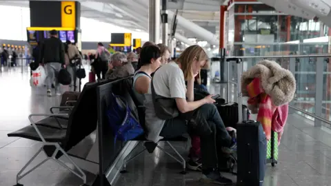 Orang -orang Reuters duduk di kursi di terminal di Bandara Heathrow. Mereka duduk dengan koper di depan mereka.