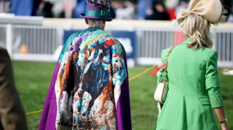 Reuters A woman in a colourful cloak which has a picture of horse racing on it, crosses the racecourse at Aintree