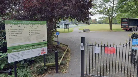 The entrance to a park. There is a black gate on the right and an opening in the path to the left of the gate for people to walk through. There is a cream and green sign that reads "Victoria Park". Beyond the gate there is a large green space and tennis court which are separated by a path.