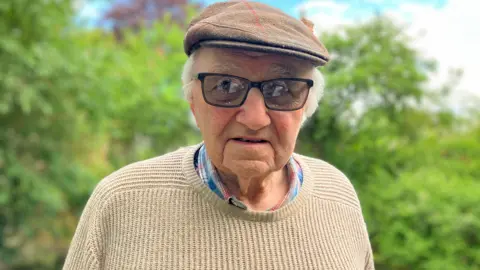 Martin Fordham looking at the camera. He is wearing a beige knitted jumper, tinted glasses and a beige flat cap. Behind him are trees and bushes, out of focus. 