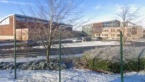 Google A modern school campus viewed from behind a metal fence. The ground is lightly covered with snow. On the left, there is a large structure with a curved roof that looks like part of a sports hall or gymnasium.
On the right, a multi‑storey, contemporary school building features large windows and a mix of brick and lighter‑coloured materials. There are a few leafless trees in the foreground.