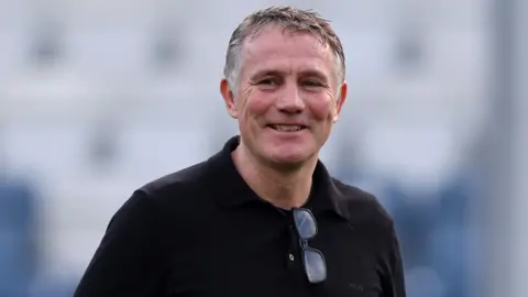 A smiling Wrexham manager Phil Parkinson wearing a black shirt with spectacles hooked over collar