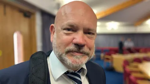 With a satchell strap over his right shoulder and white shirt, striped blue tie and blue suit, Mark Kelly poses for a photo following the council's planning meeting.