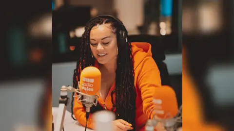 Mefus photography Aleighcia in a studio presenting. She is wearing an orange hoody, and her hair is in cornrows. She has extensions in as well. She is sat at a studio with an orange Radio Wales microphone in front of her. 