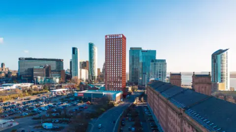 Image shows a planned 28-storey tower block to be built near Liverpool's Three Graces. The image shows the view from the Waterloo Warehouse, a long red-brick 19th century building. A number of tall buildings obscure the view of the waterfront. 
