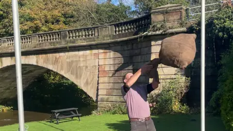 A man hurls a sack of apples in the air on a picturesque spot next to the river in the village of Alton, with an old bridge in the background