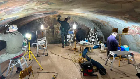 Three people, on a platformed area, in a room, with a painted ceiling above them, and painting to the end of the room. One man is standing, with an arm up, the other men are sitting down. They are all working, there are lights around them and equipment and ladders. 