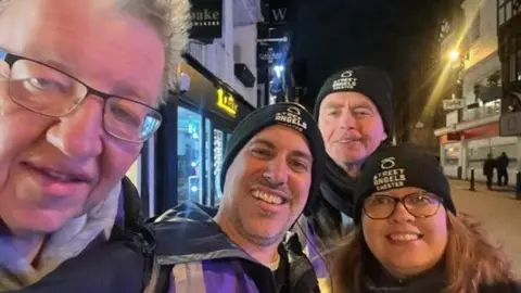 Street Angels Chester Four men and a woman, three of who are wearing black beanie hats with Street Angels Chester stitched onto the front in white lettering, smile for a selfie on a city centre street at night. 