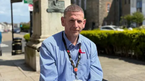 James has short greying brown hair and a beard and wears a blue shirt with a black lanyard around his neck. He sits outside Camborne Town Hall with the pillar of the statue of Richard Trevithick behind him. 