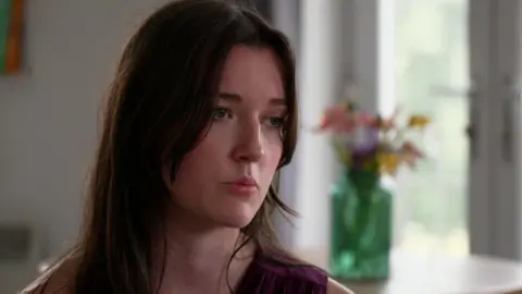Charlotte, who has long brown hair and is wearing a maroon sleeveless top, sits in a white room beside a window with a vase of flowers on the sill