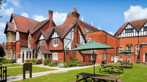 Trustees of the Natural History Museum The front exterior of a large detached or semi-detached house with a distinctive early 20th Century architectural style, but in a Tudor style. There is an open lawn in the front with tables and benches. One of the tables has a green parasol covering it.