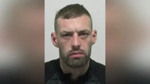 Mugshot of Burdis. He has an unhappy expression, with short dark hair and a thin dark beard and moustache.