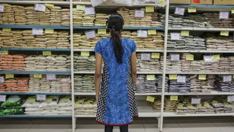 Bloomberg via Getty Images A woman wearing a blue printed top stands in front of a grocery store shelf stocked with pulses and grains