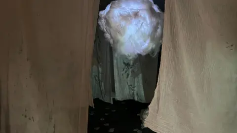 Nathan Slate Sheets are hanging from the ceiling in a small room. There is a construction made to look like a cloud in the middle, which is white and lit up.