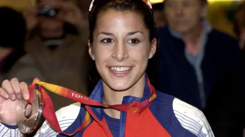 Getty Images Olympian Shelley Rudman holding her silver media in her Team GB coat and smiling. She has brown hair and eyes and there are people stood behind her.