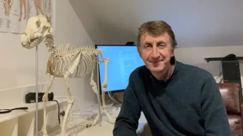 Ava Taylor Dr Sean Taylor sits at his desk next to a skeleton of a dog.