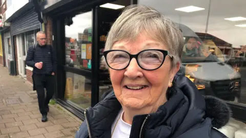 Irene Williams smiling looking at the camera. She is standing on Newport Road with cars driving behind her. She is wearing sunglasses and a black coat. 