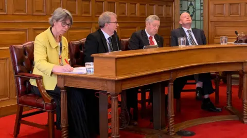 MLCs Dawn Kinnish, Paul Craine, Peter Greenhill and Bill Henderson sitting in the Legislative Council. The room has wooden panelling on the walls and a red carpet.