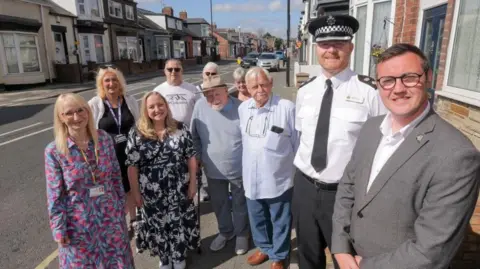 In the right is Sunderland City Council Leader Cllr Michael Mordey with Superintendent Mark Hall. From left: Deputy Leader of Sunderland City Council and Cabinet Member for Health, Wellbeing and Community Safety, Councillor Kelly Chequer, and second from left in the foreground Cabinet Member for the Environment, Councillor Lindsey Leonard with Hendon residents. They are smiling and standing on the pavement. The street behind them has rows of terraced houses and a number of parked cars.