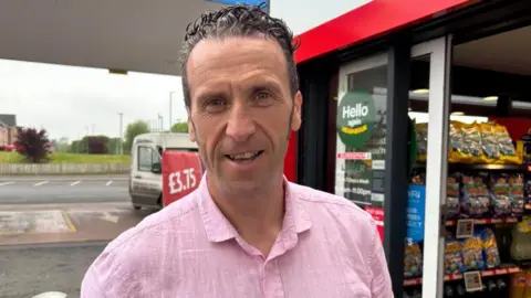A man with gelled black and grey hair smiles into the camera. He is wearing a pink short sleeved shirt. He is standing at the doorway of a shop with a garage forecourt behind him.