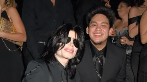 Getty Images A photograph of pop star Michael Jackson with black hair and aviator sunglasses. Pictured with Prince Azim at his 25th birthday party at Stapleford Park Hall. They are both wearing all black suits
