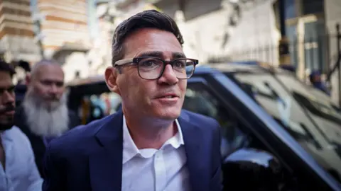 Andy Burnham wearing a dark blue suit jacket and white shirt.
