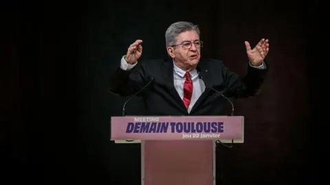 Getty Images Leader of far-left party, La France Insoumise Jean-Luc Mélenchon before a podium reading "Meeting, DEMAIN TOULOUSE (tomorrow Toulouse), Jeu 22 janvier".  He is speaking animatedly 