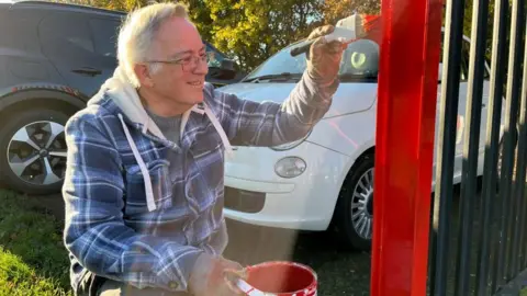 BBC A man with grey hair and glass painting a fence with red paint. There is a black car and white car behind him.