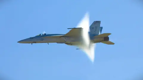 Getty Images A grey fighter jet flying across a blue sky and creating a cloud-like pressure wave towards its tail.